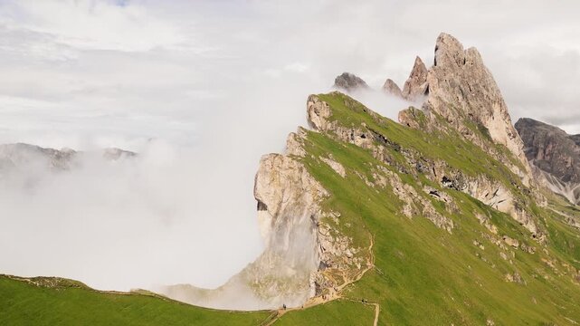 Aerial view tourist walk hike Seceda ridge with Odle peaks rise over clouds with steep limestone cliffs green alpine slopes winding hiking trail. Panoramic Dolomites landscape. South Tyrol Italy