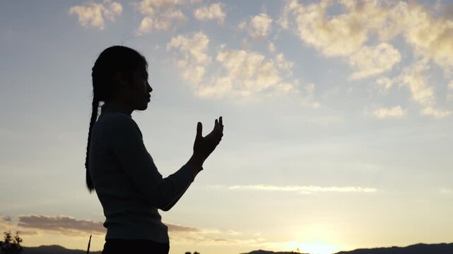 A silhouette of a woman open palm  praying on a mountaintop at sunset, 4K slow-motion video.