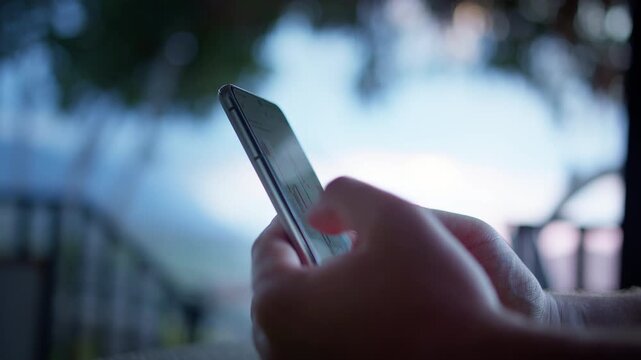 Side view close-up of man holding smartphone with stock trading application. Market charts and analytics on screen while soft daylight and blurred trees form modern financial technology mood.