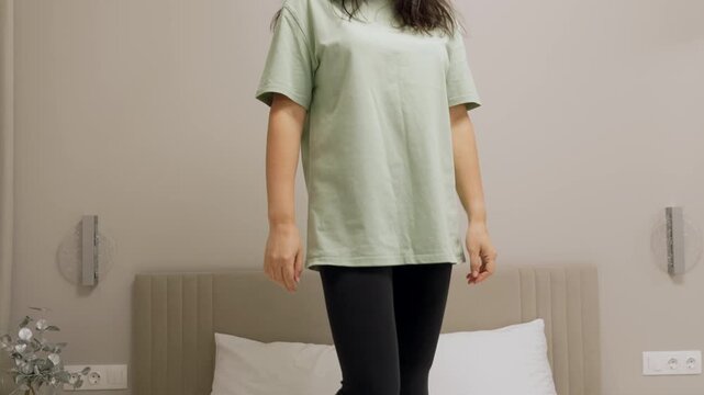 A cheerful brunette woman in casual clothes has fun dancing on her bed in a modern bedroom. She smiles, enjoying her freedom and a happy moment at home. Feeling good concept