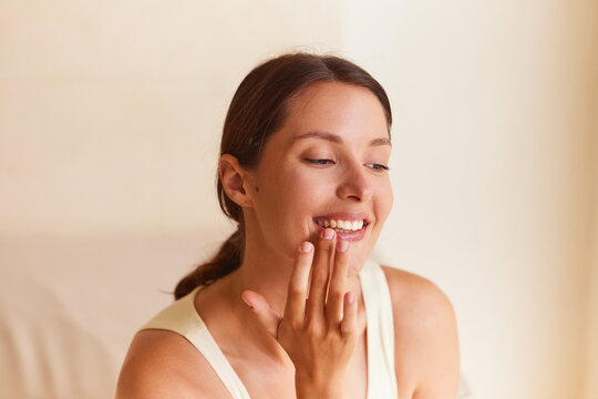 Lip oil. Woman gently pressing open hand against her lips with eyes nearly closed, serene sensory expression. Clean studio beige background, close-up beauty portrait. 