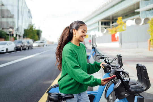 Smiling woman unlocking shared bike on city street for sustainable travel