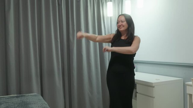 A happy middle aged woman with long dark hair in a black dress is dancing joyfully in her bedroom. She smiles, feeling carefree and positive, enjoying the moment of freedom and self expression at
