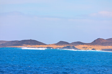 Naklejka premium Volcanic coastline of Lobos Island, near Fuerteventura, Canary Islands, where waves crash against dark rocks and sandy shores. Dormant volcano rises under a hazy sky, overlooking deep blue waters