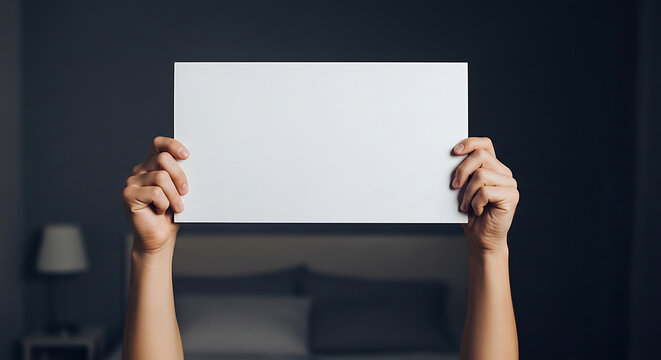 Two hands holding up a blank white rectangular sheet of paper against a blurred dark indoor background with mock