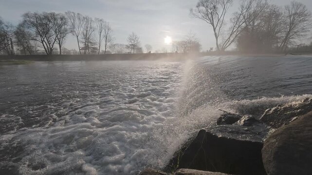 Small river weir with flowing water creating a gentle cascade. The scene captures movement, texture of water, and a calm natural atmosphere in a countryside setting.
