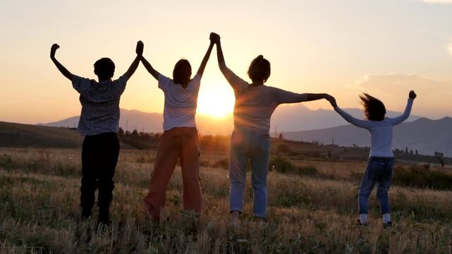 Rear view mother and three children holding hands jumping together in field at sunset looking into distance and enjoying scenic mountain sunset. Family scene outdoors. Happy family together.