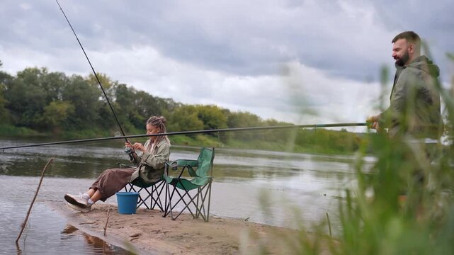 Happy young couple spending quality time together fishing rods from sandy river bank on cloudy day. Romantic outdoor activities and a shared hobby to connect with nature