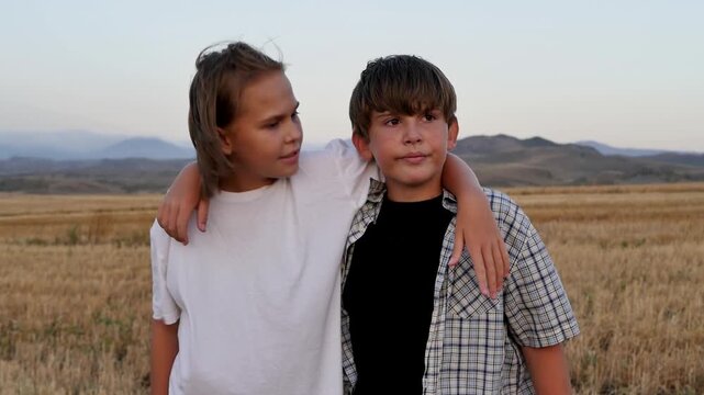Cute teen boy and girl standing in mowed rural field with arms around shoulders thinking and deciding something. Wind blowing their hair. Brother and sister caucasian kids outdoors in evening at dusk