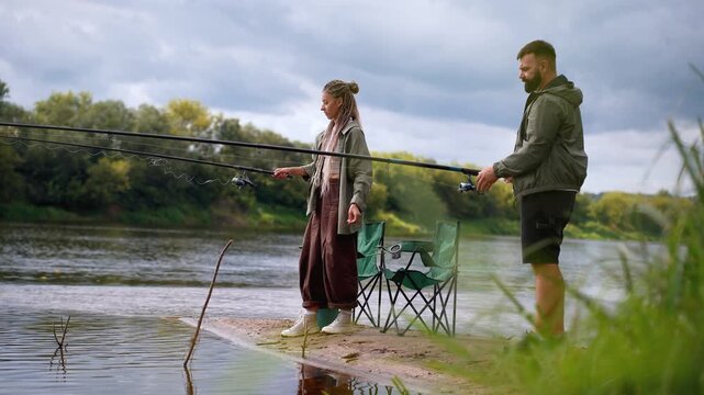 Millennial man and woman enjoying a relaxing day outdoors, standing on a sandy riverbank and preparing their fishing gear together for a recreational angling session in nature