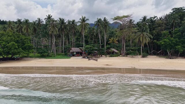 Low drone approaches sandy tropical beach from the sea revealing nipa hut cottages nestled among tall coconut palms with driftwood on shore and cloud-covered Palawan mountains behind