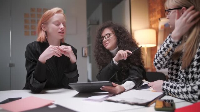 Focused female coworkers discussing ideas and working together in a modern office environment. Women, colleagues or team members gathered around a tablet and documents while sharing feedback, planning