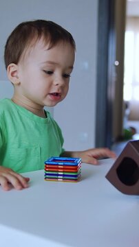Cute dark-haired boy sitting at the little desk. Baby looks at a range of toys lying on the table. Vertical video