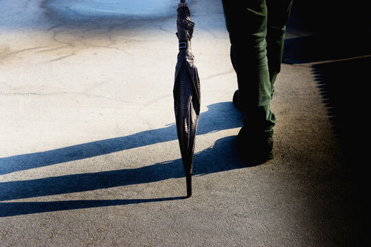 Authentic conceptual shot of a person walking with a closed umbrella on a textured asphalt ground featuring long artistic shadows under low sunlight for city lifestyle design.