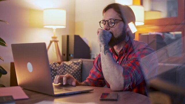 Professional man using a laptop while working in a cozy modern workspace during evening hours. Young male, businessman or specialist wearing glasses and casual plaid shirt, focused on digital tasks