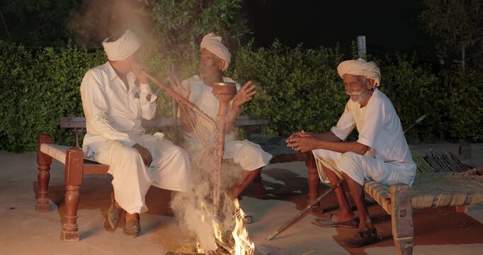 Indian old aged man wear white kurta pajama sitting on woven cot bed enjoy desi life at rural area. Happy three male group talk speak share gossip do hookah hold pipe spend night time village place
