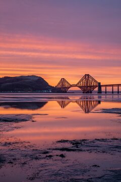 Spectacular Forth Bridge at Sunset with Vibrant Orange and Pink Sky Reflected in Calm Water and Rocky Shoreline at Dusk