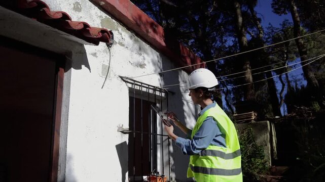 Female civil engineer with a hard hat and safety vest inspecting a building's exterior wall damage while using a tablet for data entry on a construction site