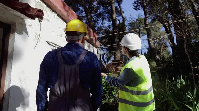 Civil engineers inspecting an old building's roof structure for damage, a female architect explaining renovation plans to a male construction worker on a tablet