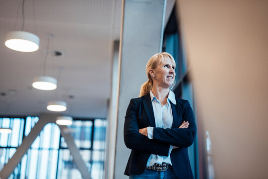 Smiling mature businesswoman standing with arms crossed near columns in corridor