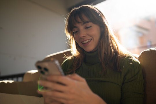 Smiling woman using smartphone on sofa in bright indoor setting