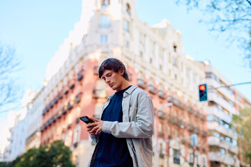 Fototapeta premium Young man standing outdoors in a city, focused on his mobile phone device, connecting with technology and urban life