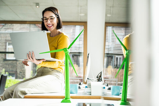 Smiling businesswoman using laptop sitting by wind turbine model on desk in office