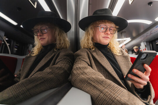 Young man in hat using smartphone on subway with reflection