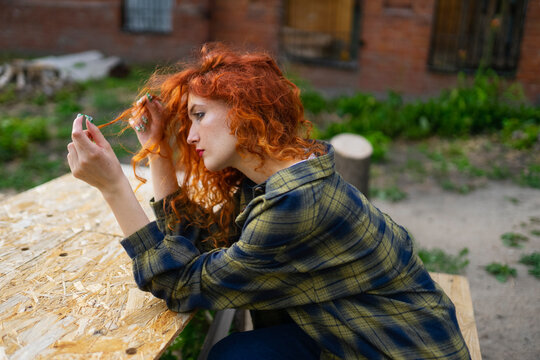 Woman with red curly hair in plaid shirt sitting outdoors by house