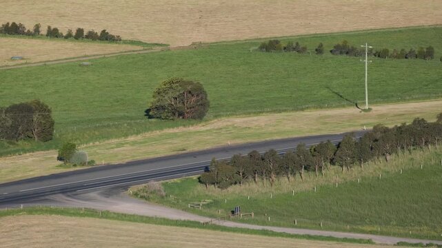 Expansive rural farmland with a country road and vehicles passing at Camperdown, Victoria, Australia. Elevated view of vast agricultural fields and traffic. Regional transport, quiet countryside