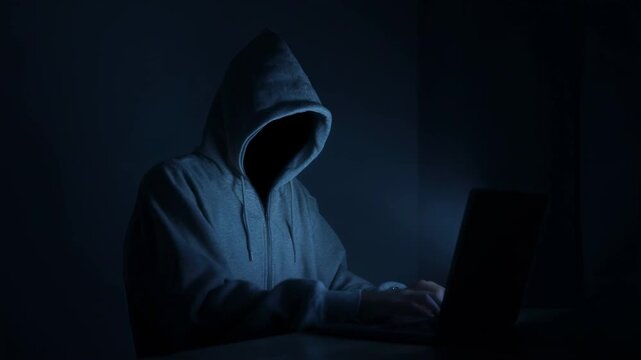 Anonymous hacker typing on laptop in dark room. Faceless cybercriminal in hoodie coding malicious software. Cyber security threat and internet fraud