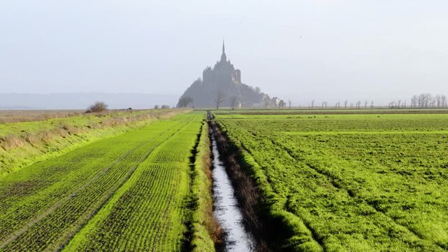 Aerial view of the historic Mont Saint-Michel abbey rising above misty fields and a narrow water canal in Mont Saint-Michel, Normandy, France.