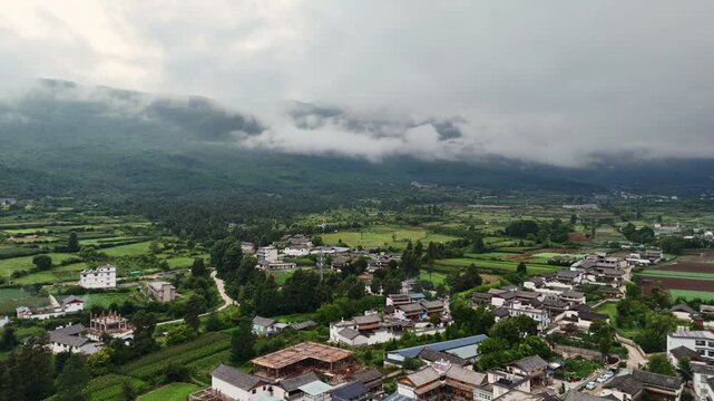 Baisha town houses farmland and mountain valley under clouds near Fuguo Monastery Yunnan, aerial tracking left