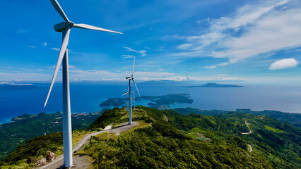 Wind turbines in the mountains. Wind turbines above tropical islands and blue ocean. Aerial view of renewable energy infrastructure on mountain ridge with scenic seascape and coastal landscape. © Houston