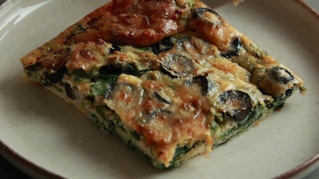 Close-up of a slice of baked egg frittata with olives and spinach on a rustic plate