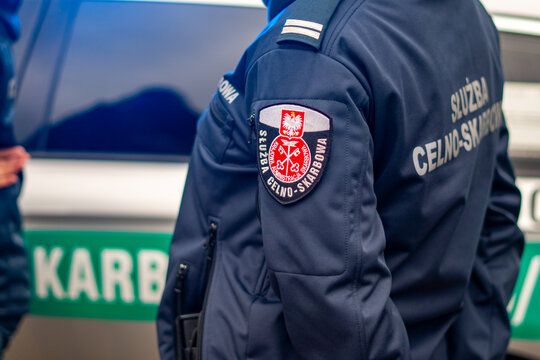 Polish Customs and Revenue Service officer in navy blue uniform with official KAS patch, customs control and tax administration enforcement, officer on duty next to a patrol vehicle.