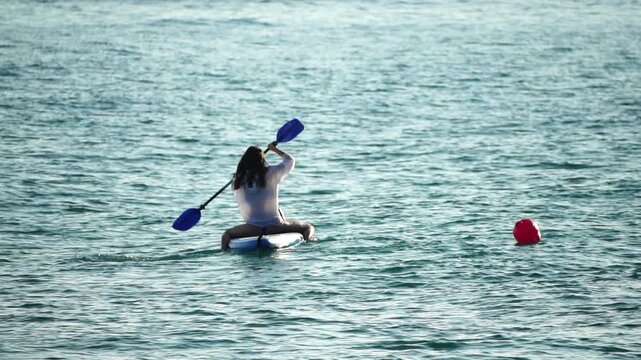 Paddleboard woman ocean woman in white shirt sitting on a paddleboard and rowing across the calm blue sea for summer fitness