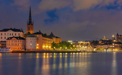 Blue hour view of Stockholm waterfront in Sweden with illuminated historic buildings, church spire and reflections on calm water.