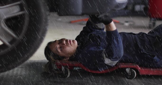 Woman auto tech lying on creeper after service, fixing wheel area while rain overlay marking parts