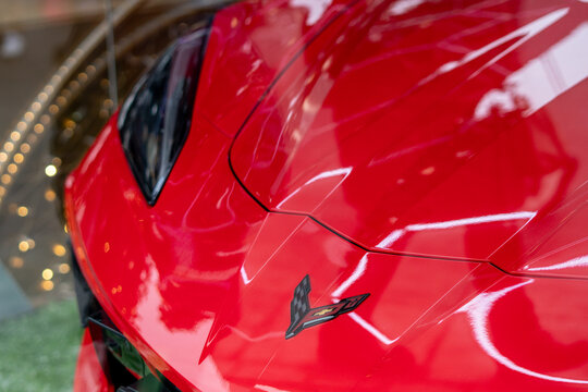 Red Chevrolet Corvette C8 hood with emblem close-up, shiny car paint and aerodynamic body details, luxury supercar on display with festive bokeh lights in the background, automotive design.