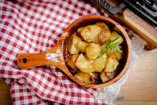 View of golden roasted potatoes nestled in a wooden bowl, sprinkled with salt and a sprig of rosemary, against a red and white checkered cloth, Pula, Croatia.