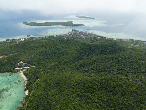 Aerial view of lush green forests meeting the turquoise sea, with the settlement nestled atop, framed by distant islands, Karimunjawa, Central Java, Indonesia.