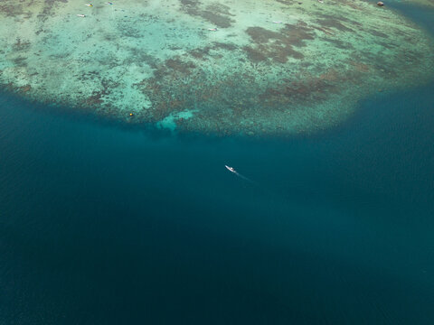 Aerial view of a solitary boat glides across the tranquil, deep blue sea, contrasting with the vibrant turquoise reef below, Karimunjawa, Central Java, Indonesia.