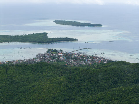 Aerial view of a dense village nestled amongst lush greenery meeting the turquoise sea, with distant islands under a soft sky, Karimunjawa, Central Java, Indonesia.
