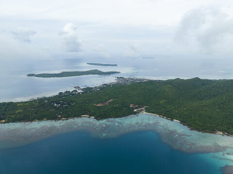 Aerial view of the lush, green island meeting the tranquil, turquoise sea under a vast, overcast sky, Karimunjawa, Central Java, Indonesia.