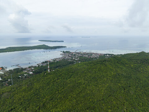 Aerial view of lush green hills cascading towards the coastal town, where the sea meets the land in a vibrant display of nature's beauty, Karimunjawa, Central Java, Indonesia.