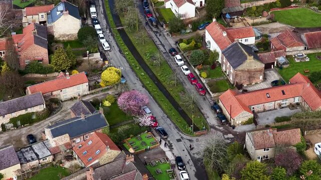 Top down aerial footage of Bishop Monkton road with a narrow water channel trees houses gardens and parked cars showing a traditional rural village street in North Yorkshire during spring time