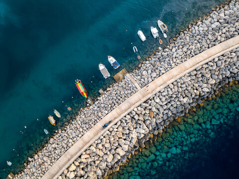 Aerial view of boats bobbing gently on the azure waters beside the rugged, rocky breakwater of Mgarr Harbour, Gozo, Malta.