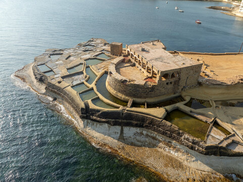 Aerial view of geometric salt pans reflecting the sky, contrasting with the rugged stone structures of Qolla l-Bajda, Iz-Zebbug, Gozo, Malta.