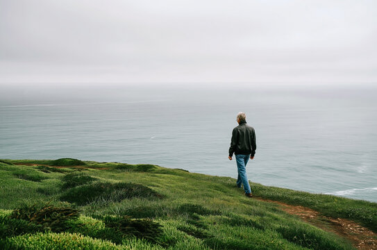 Lone Woman Walking on Green Coastal Cliff Overlooking Misty Ocean Horizon in Moody Overcast Atmospheric Landscape